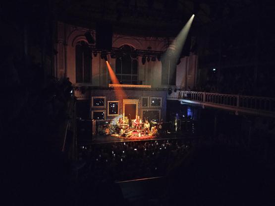 Concertfoto van Lucy Dacus in Paradiso, vanaf het balkon genomen. De band is verlicht door een rood en wit spot licht. Ze zitten met z'n allen (gitaar, viool, zang, keyboard en akoestische gitaar) op een halfrond vloerkleed voor het 'kampvuur' deel van de set. De drummer heeft even pauze :)
Lucy heeft net een pop van Nijntje cadeau gekregen en die staat vooraan.