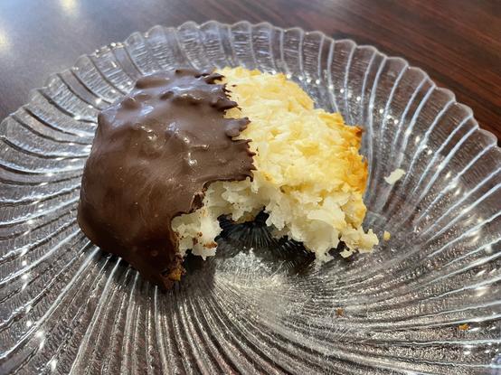 A coconut macaroon on a glass plate with a bite taken out of it. Half of the cookie has been dipped in chocolate so it resembles a traditional 'black and white cookie.'