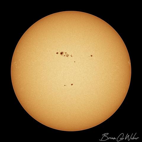 A full disk monochrome image of the sun colorized to orange with a thick cluster of sunspots on the upper middle part of the surface.