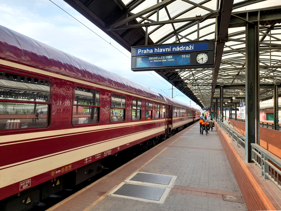 A red train at Prague main station. An electronic sign indicates the train is going to Brussels via Amsterdam. Further away two people are walking on the platform with their bikes.