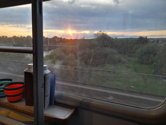 A view from the train window as the sun is coming up in between the clouds behinds some trees. On a narrow table in front of the window are two water bottles, two cups and a train ticket. To the left part of the ladder to get to the top bunks is visible.