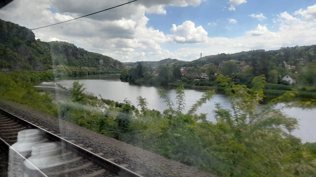 A view from the train window that shows a curve in a river with low mountains in the distance. On the opposite side of the river, to the right, houses are visible in between the trees. In the bottom left corner train rails are visible and in a reflexion also two water bottles.
