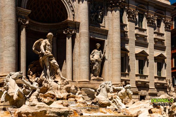 Close daytime view of Trevi Fountain in Rome showing central statue of Oceanus, flanking figure with spear, winged horses, and tritons amid flowing water against ornate Baroque columns and windows.