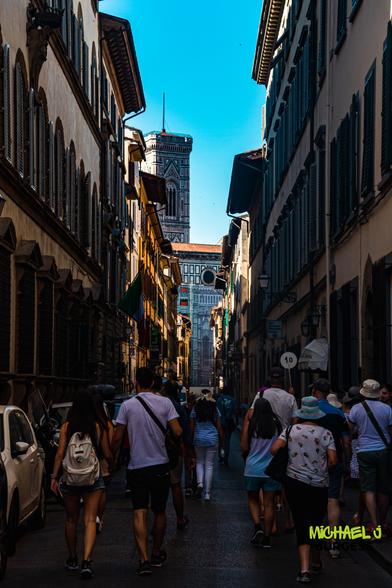 Vertical photo of a narrow street in Florence filled with tourists walking toward the distant Duomo; tall shuttered buildings cast shadows while the ornate bell tower and cathedral façade rise against a clear blue sky at the end of the lane.