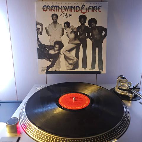 A vinyl record sits on a turntable. Behind the turntable, a vinyl album outer sleeve is displayed. The front cover shows 5 members of Earth, WInd, and Fire. 4 are standing and posing for the camera, while the 5th is parallel to the ground doing a dance move. They are all dressed in '70s soul/funk attire