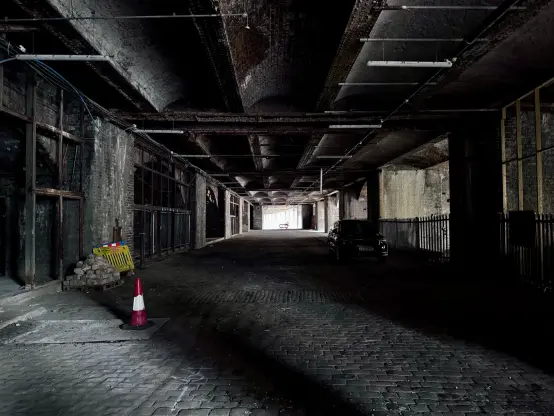 Entrance to the site. A long corridor with sunlight coming in from the right. It is a wide space with cobblestones and an arched roof. There are arches to the left, which are partially blocked off. The whole space is rather dark and dank.