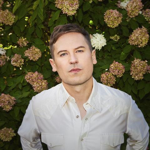 A white man with neatly trimmed auburn hair looks directly into the camera. He wears a white Western-themed shirt and stands against a large hedge with white flowers.