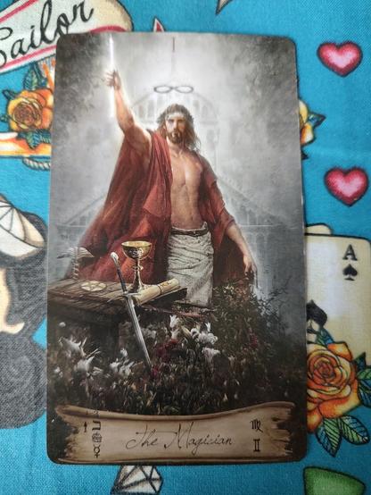 A man in white and red robes, he holds a wand up in his right hand. In front of him a table with other tools including a chalice (cup), sword, and a pentacle. There are flowers in front.