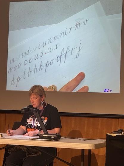 A person is seated at a table with a microphone, demonstrating English Roundhand calligraphy. In the background, a projection displays handwritten letters on paper. The individual appears focused on their writing.