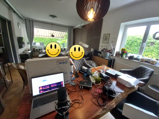 a living room with a podcast recording setup.  latop and microphone in the front, 2 people made anonymous in the background.  the room has large windows overlooking a green garden.