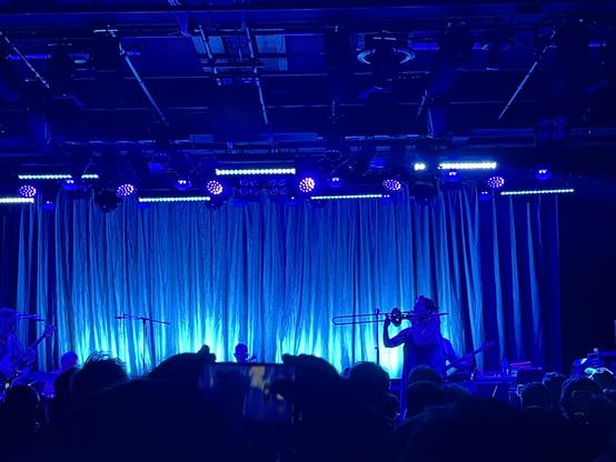The band’s vocal lead Laetitia Sadier playing a trombone on stage set in blue light gradients.