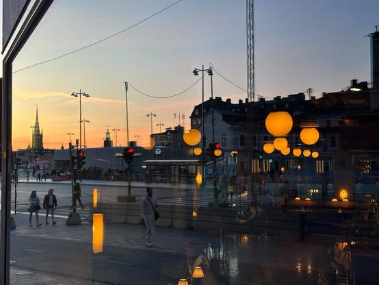 a reflection of the sun setting over an urban square in a large plate glass window. In the dark areas, where buildings are reflected, clusters of spherical lamps shine through from the interior