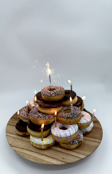 Doughnuts in different colors & flavors stacked on top of each other like a little pyramid with lit candles inserted here & there - and on top sits a sparkler.