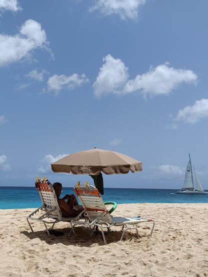 Gerardo, nude, proofreading a soon-to-be-published naturist fiction book, on a beach chair and under a beach umbrella on Cupecoy Beach, Sint Maarten, Netherlands Antilles.  There is a catamaran sailing to the west in the background.