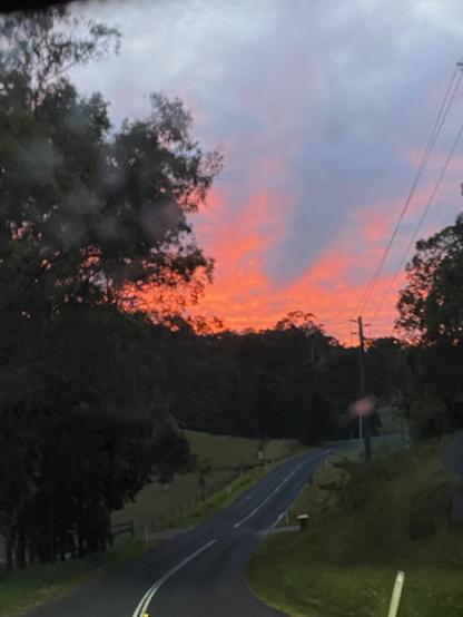 A view down a road that is going down hill. Above is an overcast sky but in the horizon the clouds are very pink, with pink tendrils upwards.