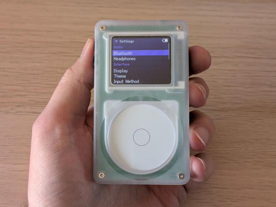 A photo of a portable media player held in a hand. It's a rectangular device with an LCD screen and a circular control wheel. On the screen is the settings menu, showing options for Bluetooth, Headphones, Display, Theme and Input Method.