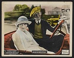 A man and a woman are sitting in a convertible car. The woman is wearing a sleeveless, white dress and a white hat. The man is wearing a black suit, a black tie, a white shirt, a yellow vest and a yellow cap.