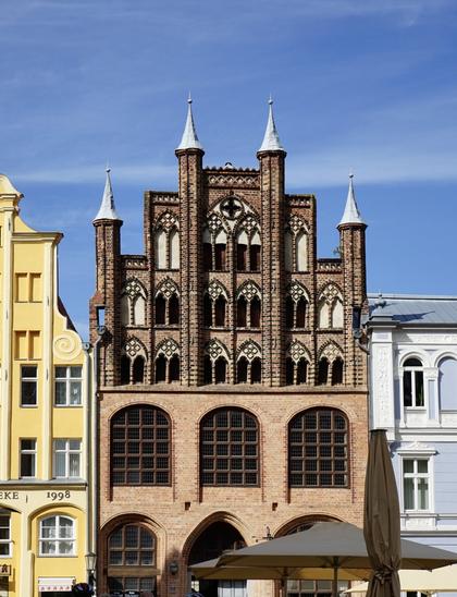 Photo d’une vieille maison bourgeoise en brique et à pignon à gradins typique d’Allemagne du Nord. De petites fenêtres en ogive à meneaux viennent aérer le pignon, dont la forme est soulignée par de fausses tourelles décoratives (je crois que c’est ce qu’on appelle des pinacles). En dessous du pignon, on a une partie qui a visiblement été refaite, avec trois grandes fenêtres en arc qui occupent la largeur de la façade. On voit mal le rez-de-chaussée, également refait, comportant apparemment encore deux grandes fenêtres et une grande porte à voussures. La façade se trouve entre deux autres vieilles maisons qu’on aperçoit un peu sur les côtés, l’une crépie en jaune, l’autre en bleu pâle et blanc.