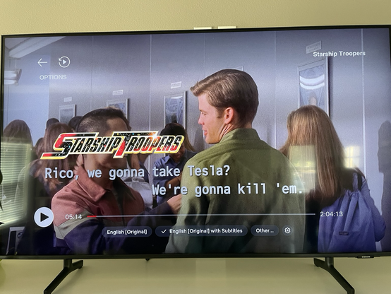 Screen capture of Starship Troopers with closed captions. People in the hallway of a school. One person says “Rico, we gonna take Tesla?” The other responds “We’re gonna kill ‘em”