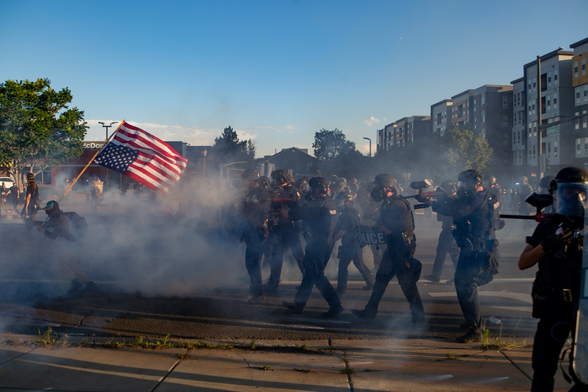 Protestor carrying a pole with the American flag inverted chased by riot police as tear gas and pepper balls fill the air.