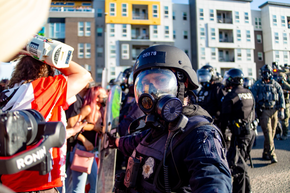 A Denver police officer in riot gear looks at the camera as protestors stand toe to toe with officers.