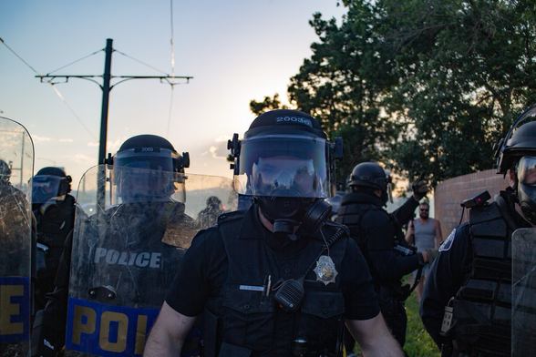 Denver Police, Colorado State Patrol, and Denver County Sheriff Deputies stand in front of the camera with riot shields at the ready.