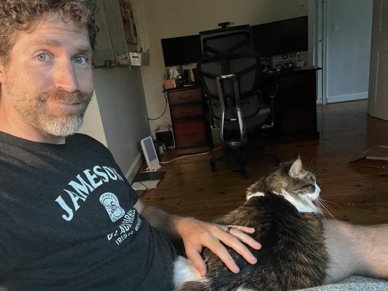 A person with curly hair and a beard is sitting on a couch, smiling slightly. A cat rests on their lap, with a hand gently stroking the cat. The background includes a desk with a computer setup and a wall-mounted board.