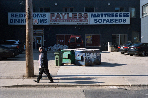 A couple walk past a wide, recessed storefront emblazoned with signs reading “Payless Mattresses & Furniture”.