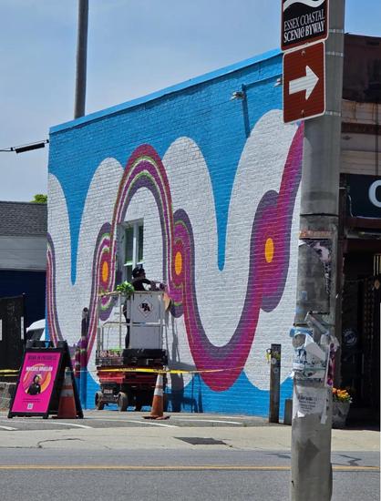 A mural on the side of a buildings it’s white and magenta circles/stripes on a blue background. In front of it is someone on a life continuing to paint.