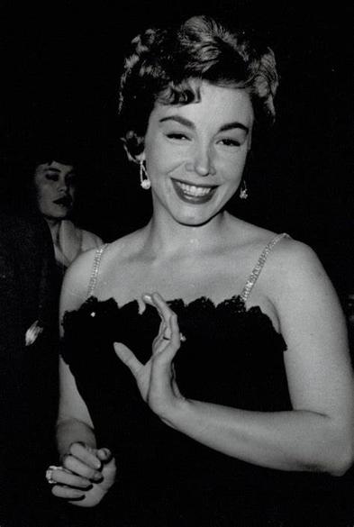 Evangelina Elizondo sonriendo y posando con un vestido negro en un evento, con un fondo que sugiere un ambiente de celebración.