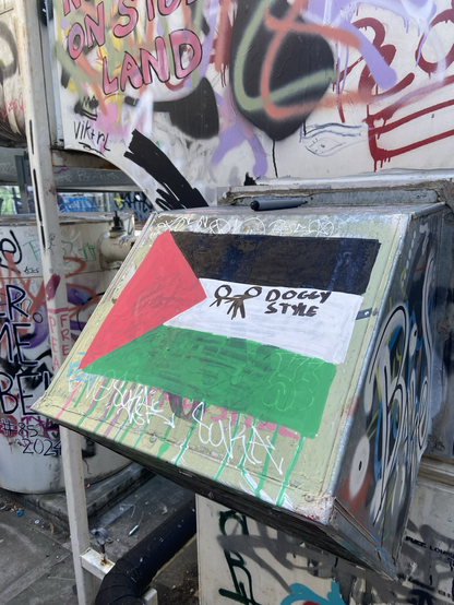 a Palestinian flag graffitied onto hvac services. another person has graffitied two small sticks figures doing “doggy style” over the flag