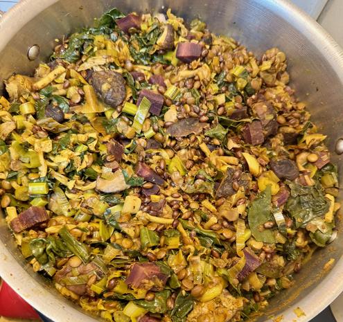 A pot of stew with an overal greenish orange appearance with large pieces of brown mushrooms scated in it. Lentils and grains of barley can be seen, along with kale leaves.