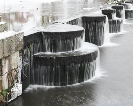 Cylindrical stone pillars form a cascading waterfall over a stepped dam. Water flows smoothly over the edges of the dark stone, creating a layered effect. The water below is calm, reflecting the overcast sky. Snow and ice partially cover the surrounding surfaces, indicating cold weather.