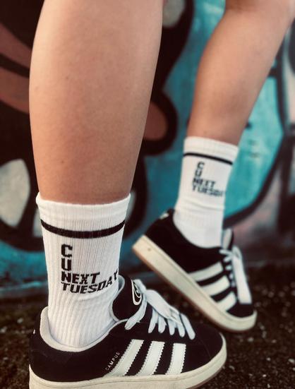 A white person's legs.  She is wearing black and white casual canvass shoes and white socks that are pulled up about a third of the way up her calf.  There is a black stripe at the top of the socks and writing that reads:

C
U
Next
Tuesday