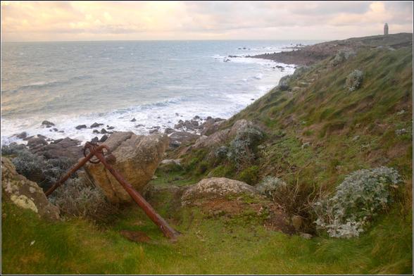 Un rivage verdoyant.
Au premier plan, une ancre abandonnée.
Au loin, un phare.