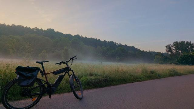 Das Bild zeigt eine Landschaftsszene bei Sonnenaufgang. Im Vordergrund steht ein E-Bike am Straßenrand. Das Fahrrad ist auf einem asphaltierten Weg abgestellt, der durch eine ruhige, ländliche Gegend führt.
Im Hintergrund erstreckt sich eine weite Wiese, über der ein feiner Bodennebel lieg. Dahinter beginnt ein dichter, grüner Wald, dessen Baumkronen sanft vom Licht angestrahlt werden. Der Himmel zeigt zarte Blau- und Rosatöne, typisch für die frühen Morgenstunden.