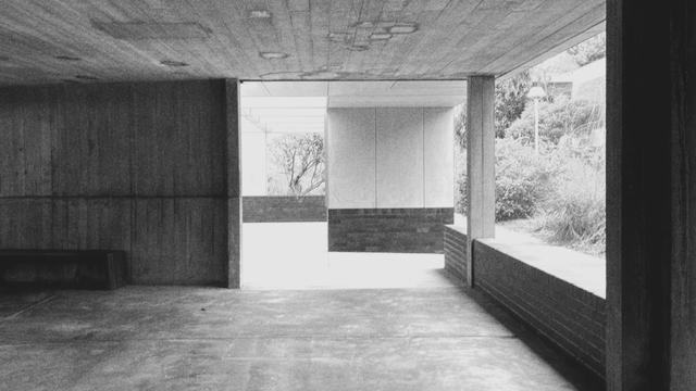 A building of concrete with openings ahead and on the right showing plants beyond. To the left is a dark wall.