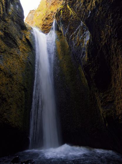 A waterfall cascades down a rocky cliff, surrounded by moss-covered stones and cliffs in a canyon. The water flows into a pool at the bottom.