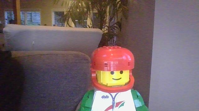A lego minifigure, scaled up, made from Lego. They are wearing a red helmet and a white jumper with green arms. On the right chest are two crossed checkered flags. In the center is a zipper that is a bit open, crossing a green and red swoosh. The background is largely grey.