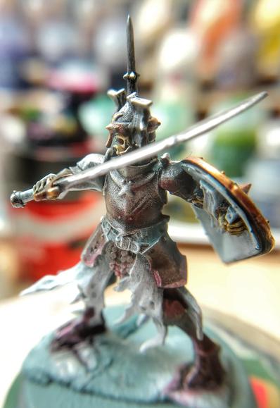 Gravelord Bone Warden miniature halfway painted. The metals are nearly done and a slight pinkish light comes from below. Seen from front.