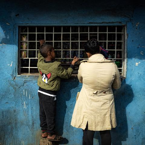 Un enfant debout sur un pavé, et un adulte, devant la grille d'une épicerie.