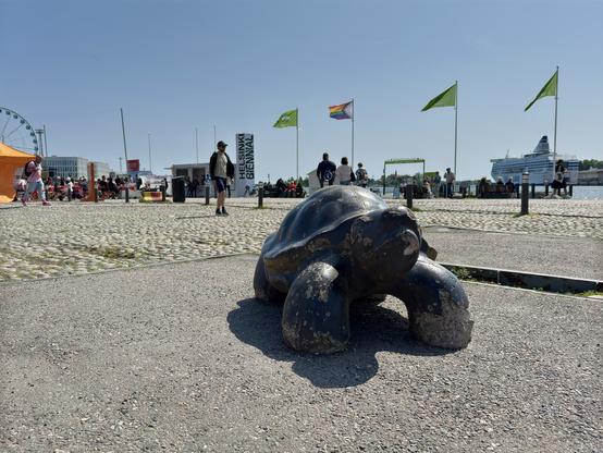 A bronze turtle sculpture is placed on a paved area with people walking in the background. There are flags flying, including a pride flag, and a ship is visible in the distance. The scene is lively, depicting a sunny day at an outdoor event.