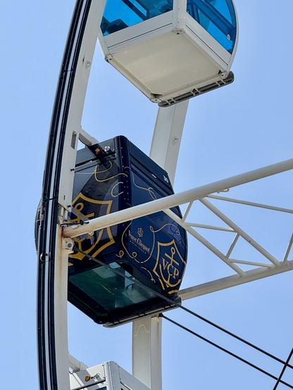 The image features a close-up of a colorful Ferris wheel gondola decorated with a logo, possibly branded by a champagne company. The background is a clear blue sky, with several gondolas visible in the frame.
