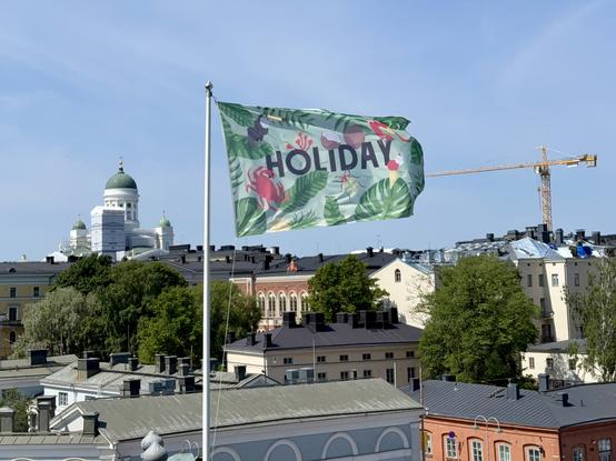 A colorful flag with the word "HOLIDAY" and tropical designs is displayed prominently against a backdrop of buildings and a church dome in Helsinki. In the distance, a construction crane is visible under a clear blue sky.