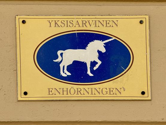 A decorative sign with a yellow background features a silhouette of a unicorn in white, centered within an oval blue area. The text reads "YKSISARVINEN" at the top and "ENHÖRNINGEN" at the bottom.