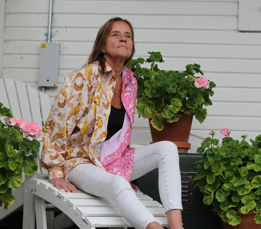 A woman wearing a two coloured shirt, with white trousers and a black undershirt. She is sitting on a white chair, against a white wall, surrounded by geraniums.