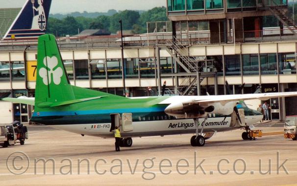 Side view of a high-winged, twin propellor-engined parked facing to the right.
The plane is mostly various shades of green, with bright green at the top, darker blue-grey stripes along the middle of the body, and white at the bottom.
There are black "Aer Lingus Commuter" titles on the lower forward fuselage, and a large white shamrock on the green tail.
There are doors open at both the front and the rear, with a man wearing a yellow hi-viz jacket leaning on the cabin door while standing outside the rear door.
Grey concrete apron fills the foreground, while a heavily-glazed terminal building stretches across the frame in the background, with the dark blue tail of a larger airliner rearing over the top of the building on the other side, with trees and grey sky in the distance.