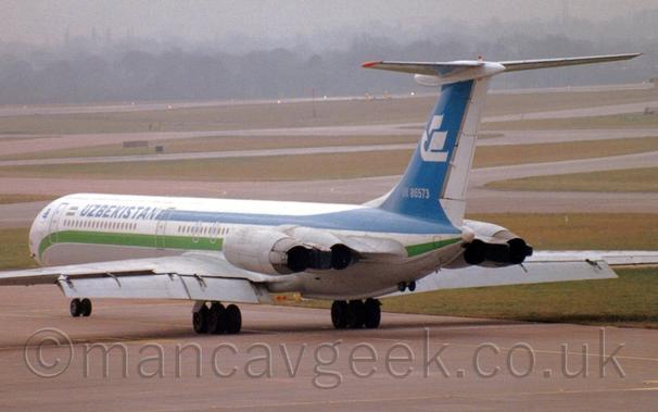 Rear side view of a 4 engined jet airliner taxiing from right to left.
The plane is mostly white, with green and blue stripes running along the body, and blue "Uzbekistan" titles on the upper forward fuselage.
The tail is blue, with a stylised white flying bird in the middle, and the white registration "UK-86573" at the base.
The 4 engines are mounted in pairs on the sides of the rear fuselage, under the tail.
Flaps are already extended from the rear of the wings, even though it has a long way to go before it gets to the runway.
Grey concrete apron fills the foreground, while taxiways wend their way through large grassed areas in the background, leading up to trees vanishing into haze in the distance, under grim grey sky.