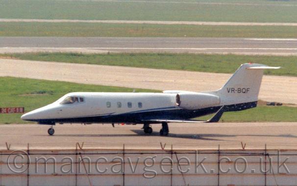 Side view of a twin engined BizJet taxiing from right to left.
The plane is mostly white, with a dark blue belly and up-turned wingtips.
The black registration "VR-BQF" is on the base of the white tail.
White hoardings tipped with black barbed wire stretch across the foreground, with taxiways and a black runway dividing a large grassed area in the background.