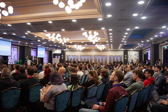 Conference audience in a big ballroom, all seated and looking towards the left, with a speaker on stage. She stands behind a lectern between two screens